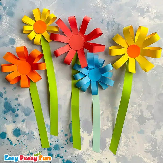 Red, blue, yellow and orange kids' craft paper flowers crafted using colorful construction paper loops. Displayed on a marbled blue and white background.
