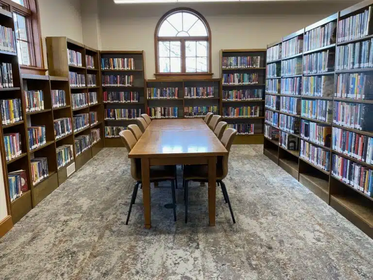 Wooden library table with chairs surrounded by shelves full of books, with an arched window behind the table.