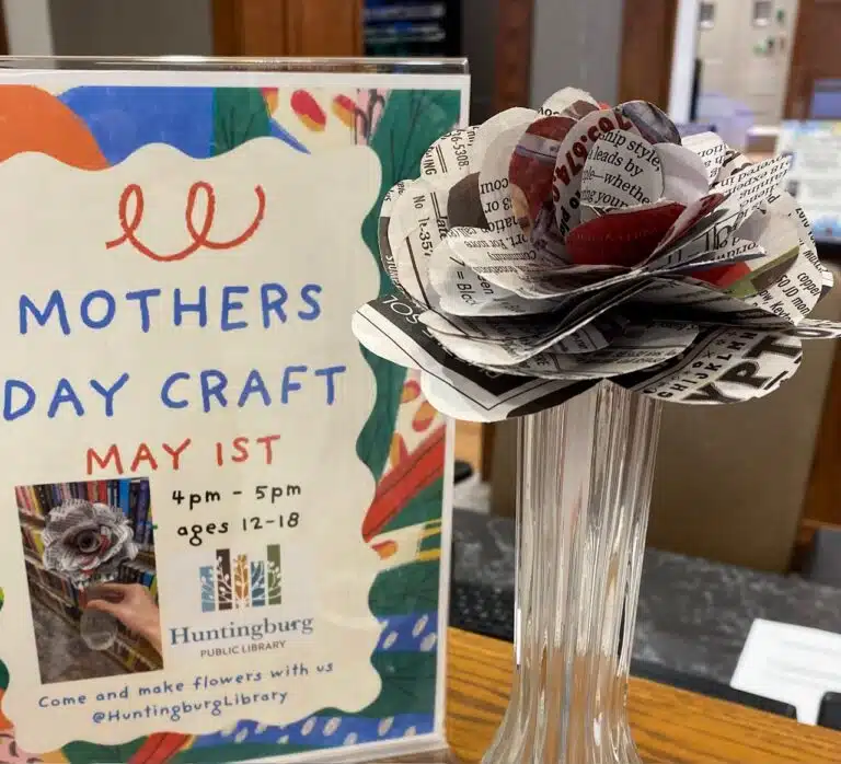 Paper flower craft made from layers of newprint, in a glass vase set next to a paper flier that says "Mothers Day Craft" May 1st.