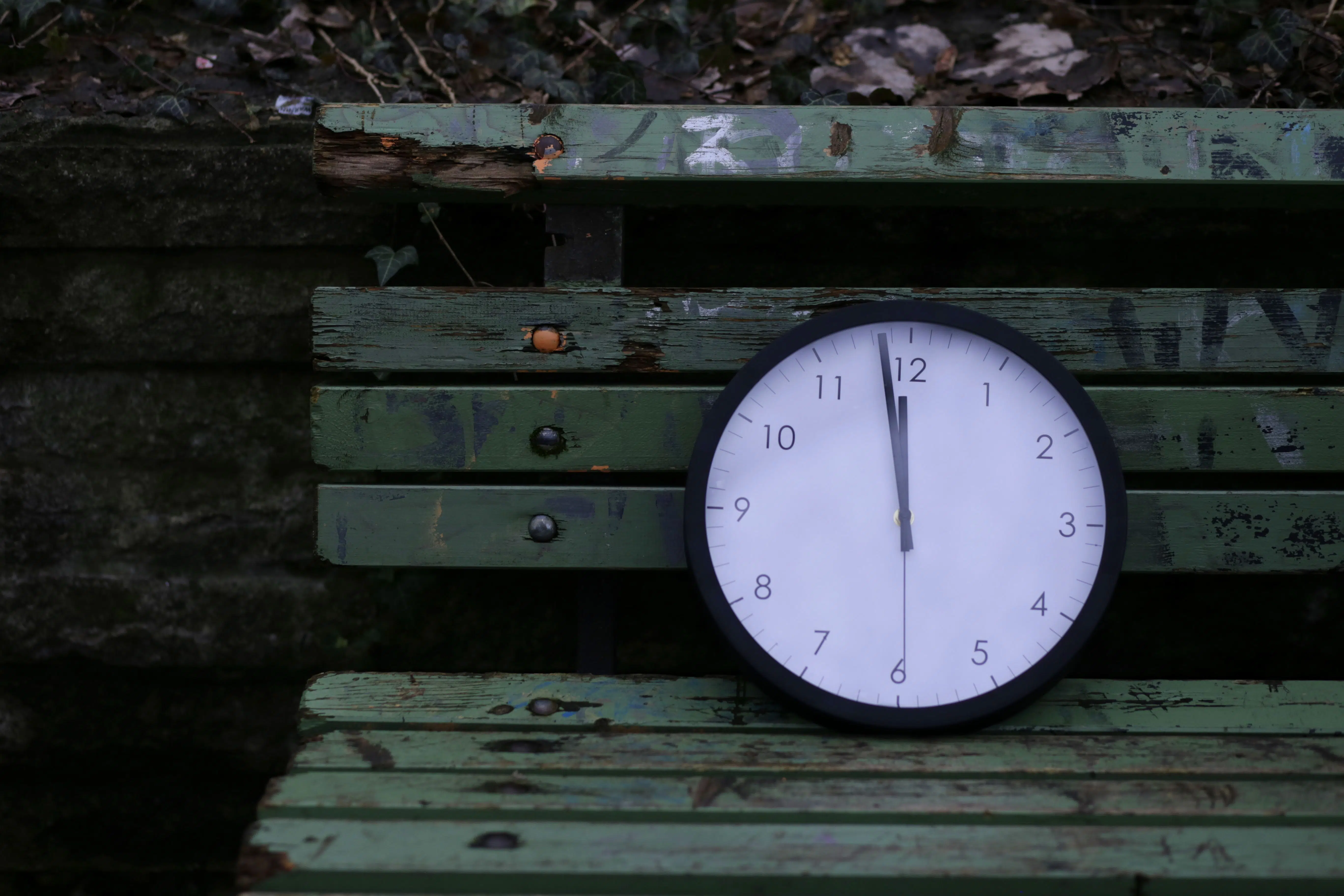 Wall Clock set to 12 noon propped up on green park bench.